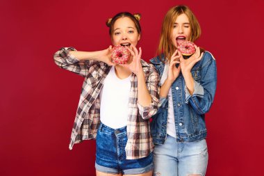 Trendy yaz kareli gömlek giysilerle poz iki güzel gülümseyen hippi kız. Kırmızı zemin üzerine izole kadın. Olumlu modelleri ile toz şeker keyfini çıkarabilirsiniz taze pembe çörek holding