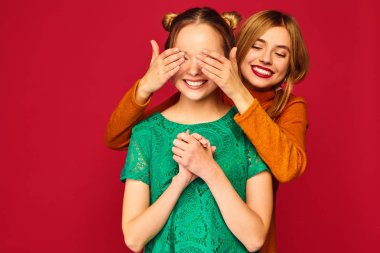 İki güzel sarışın Bayan izole kırmızı arka plan üzerinde duran fotoğrafı. Onun arkadaşı elleriyle gözlerini kapsayan kız gülümsüyor. Sarılma pozitif kadın modelleri 