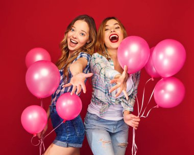 İki hipster giysili güzel kadın gülümsüyor. Kızlar kırmızı zemin üzerine poz. Pembe balonlu modelleri. Eğleniyor, kutlama Doğum günü veya tatil parti için hazır