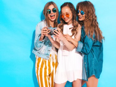 Üç genç güzel gülümseyen hippi kız trendy yaz casual elbise ve güneş gözlüğü. Seksi kaygısız kadın mavi duvara yakın poz. Retro kamera resim çekme