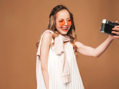 Fotoğraf selfie ile ilham alarak ve beyaz elbise giyen neşeli genç kadın portresi. Kız holding retro kamera. Model altın arka plan üzerinde poz
