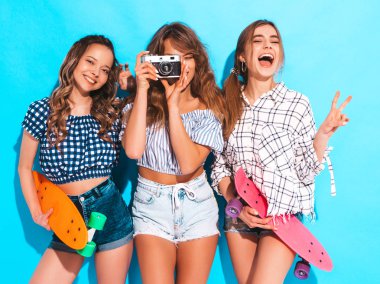 Üç seksi güzel şık gülümseyen kız renkli kuruş kaykaylar ile. Kadınlar yaz hipster kareli gömlek giysileri mavi duvara yakın poz. Retro kamera resim çekme modelleri