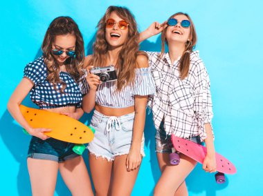 Üç seksi güzel şık gülümseyen kız renkli kuruş kaykaylar ile. Kadınlar yaz hipster kareli gömlek giysileri mavi duvara yakın poz. Retro kamera resim çekme modelleri