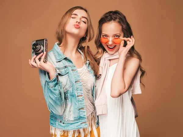 两个年轻美丽的微笑的嬉皮士女孩在时尚夏季休闲服和太阳镜 性感无忧无虑的女人摆在金色的墙附近 在复古相机上拍照 — 图库照片