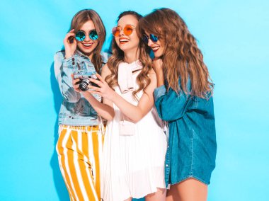 Üç genç güzel gülümseyen hippi kız trendy yaz casual elbise ve güneş gözlüğü. Seksi kaygısız kadın mavi duvara yakın poz. Retro kamera resim çekme