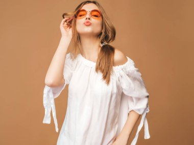 Güzel gülümseyen şirin model pembe dudakları ile portresi. Yaz beyaz hipster elbiseli kız. Model altın arka planda güneş gözlüğü üzerinde poz. Hava öpücük vererek