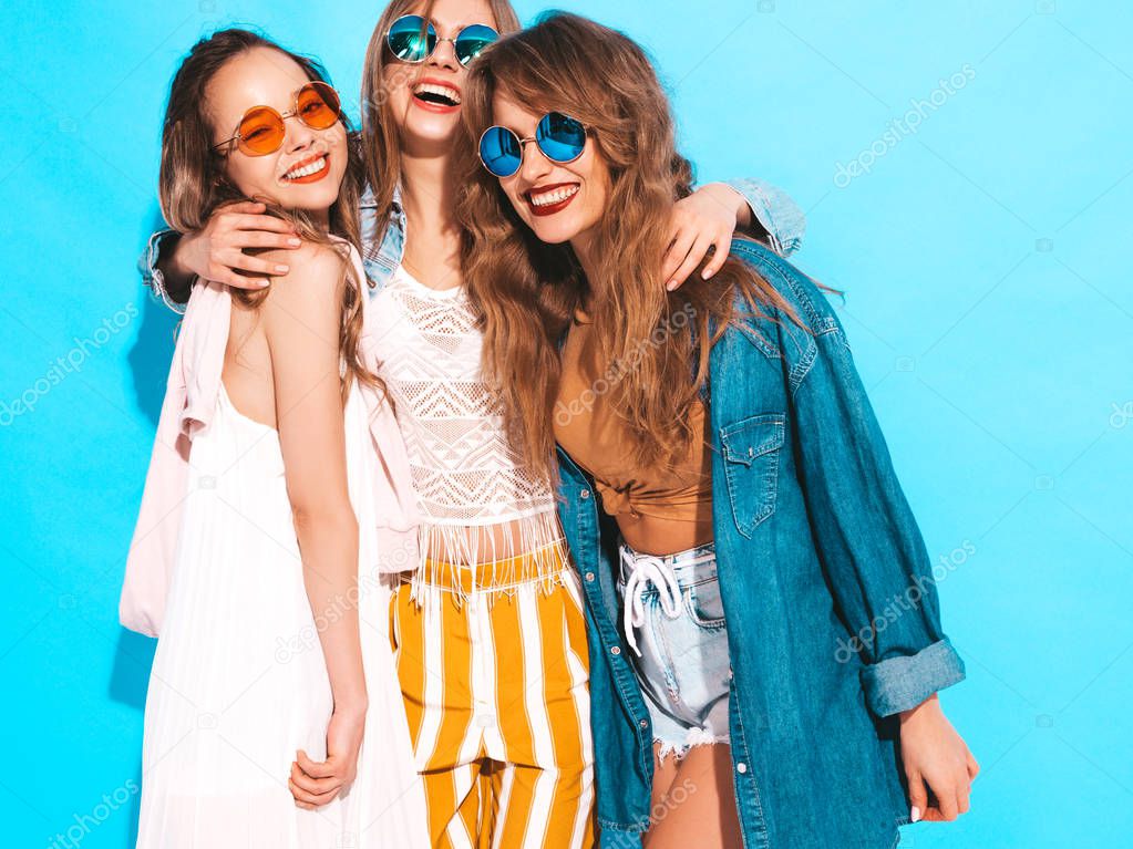 Three young beautiful smiling hipster girls in trendy summer casual jeans clothes. Sexy carefree women posing near blue wall. Positive models going crazy in round sunglasses