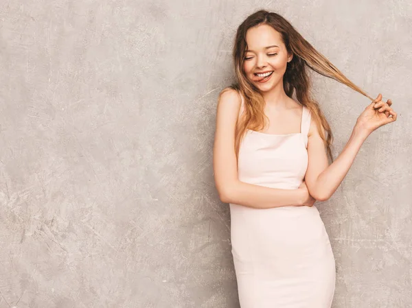 Trendy yaz hafif pembe elbiseli genç güzel gülümseyen hipster kız portresi. Seksi kaygısız kadın gri duvara yakın poz. Olumlu model eğleniyor