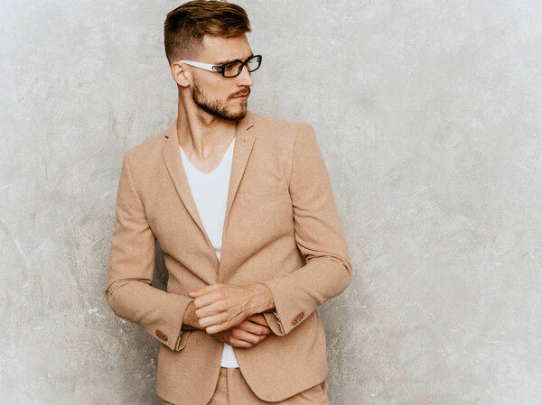 Portrait of handsome smiling hipster lumbersexual businessman model wearing casual beige suit. Fashion stylish man posing against gray wall in spectacles