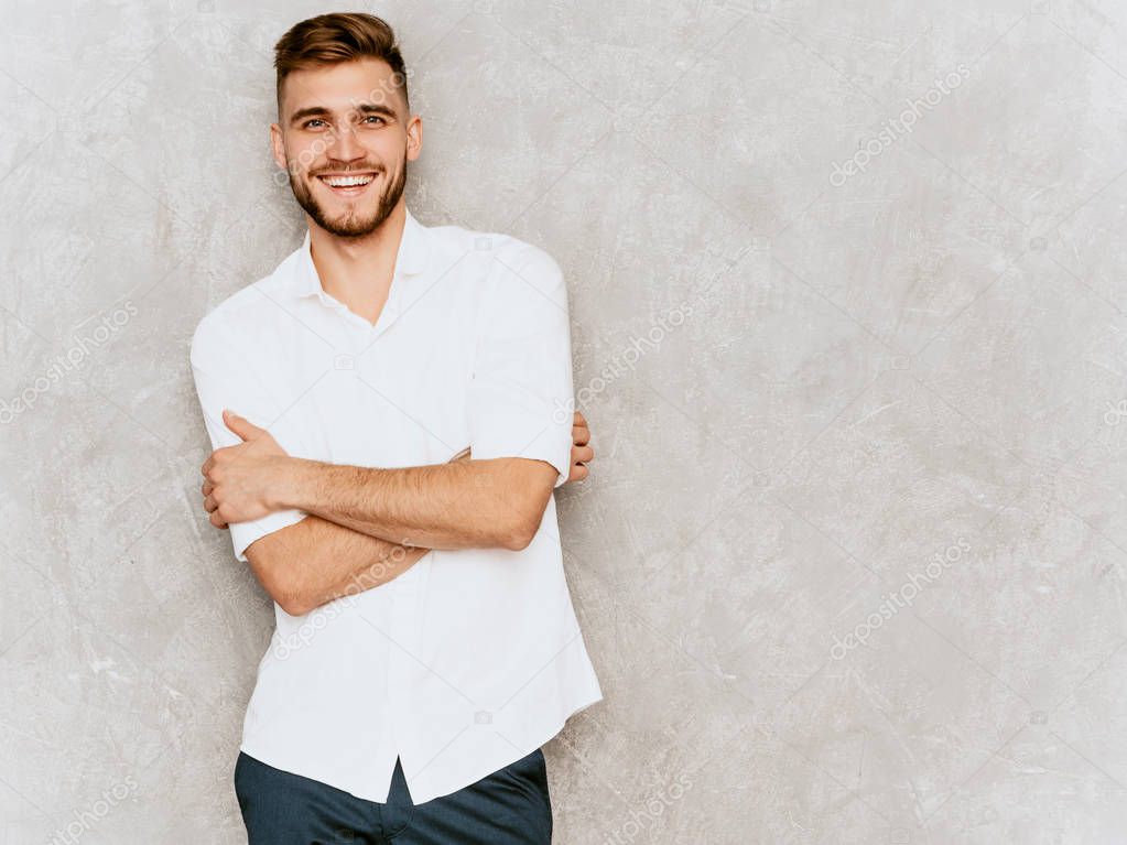 Portrait of handsome smiling hipster lumbersexual businessman model wearing casual summer white shirt. Fashion stylish man posing against gray wall.Crossed arms