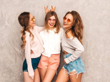Üç genç güzel gülümseyen hippi kız trendy yaz spor giysileri. Seksi kaygısız kadın gri duvara yakın poz. Eğleniyor yuvarlak güneş gözlüğü modellerinde. Parmaklarıyla kafasındaki boynuzları yapar
