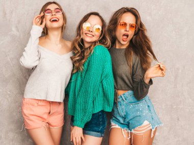 Üç genç gülümseyen güzel kızlar trendy yaz hipster giysileri güzel. Seksi kaygısız kadın gri duvara yakın poz. Olumlu modelleri içinde yuvarlak güneş gözlüğü eğleniyor