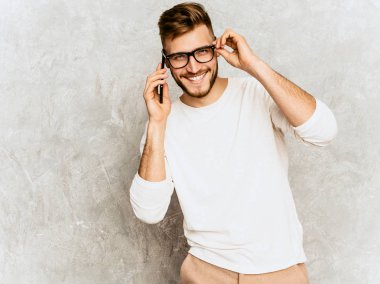 Yakışıklı gülümseyen hipster lumbersexual işadamı modeli rahat yaz beyaz elbise giyen portresi. Moda şık adam gri duvara poz. Hareket eden telefon içinde gözlük konuşma