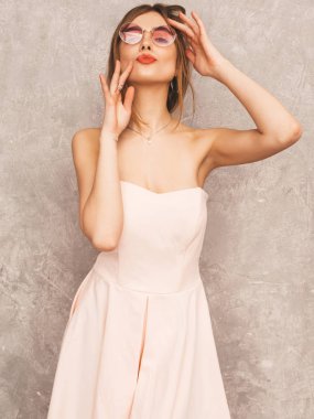 Trendy yaz hafif pembe elbiseli genç güzel gülümseyen hipster kız portresi. Seksi kaygısız kadın gri duvara yakın poz. Olumlu model eğleniyor