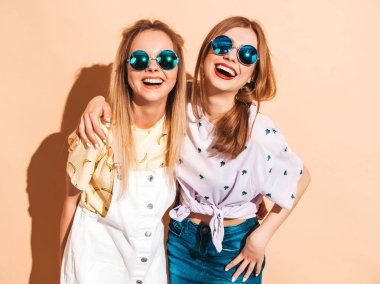 İki genç trendy sarışın hipster kızlarda gülümseyen güzel renkli tişört elbise yaz. Seksi kaygısız kadın yuvarlak güneş gözlüğü bej duvarının yakınında poz. Olumlu modelleri eğleniyor