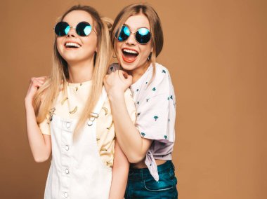 İki genç trendy sarışın hipster kızlarda gülümseyen güzel renkli tişört elbise yaz. Seksi kaygısız kadın yuvarlak güneş gözlüğü bej renkli arka plan üzerinde poz. Olumlu modelleri eğleniyor
