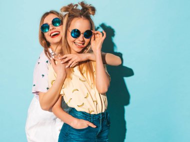 İki genç trendy sarışın hipster kızlarda gülümseyen güzel renkli tişört elbise yaz. Seksi kaygısız kadın yuvarlak güneş gözlüğü mavi duvarının yakınında poz. Eğleniyor ve dilini gösteren pozitif modelleri
