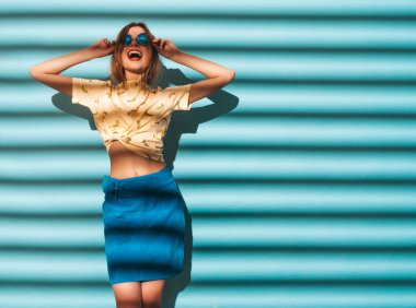 Genç güzel kadın kameraya bakıyor. Trendy kız rahat yaz sarı tişört giysileri. Olumlu erkek yüz duygular gösterir. Komik model mavi duvara yakın poz