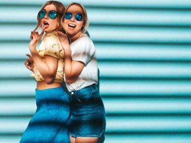 İki genç trendy sarışın hipster kızlarda gülümseyen güzel renkli tişört elbise yaz. Seksi kaygısız kadın yuvarlak güneş gözlüğü mavi duvarının yakınında poz. Eğleniyor ve dilini gösteren pozitif modelleri