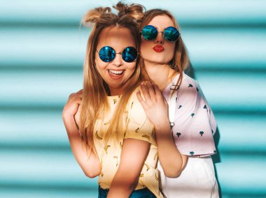 İki genç trendy sarışın hipster kızlarda gülümseyen güzel renkli tişört elbise yaz. Seksi kaygısız kadın yuvarlak güneş gözlüğü mavi duvarının yakınında poz. Olumlu modelleri eğleniyor