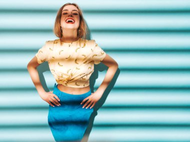 Genç güzel kadın kameraya bakıyor. Trendy kız rahat yaz sarı tişört giysileri. Olumlu erkek yüz duygular gösterir. Komik model mavi duvara yakın poz