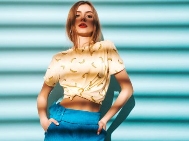 Genç güzel kadın kameraya bakıyor. Trendy kız rahat yaz sarı tişört giysileri. Model mavi duvara yakın poz