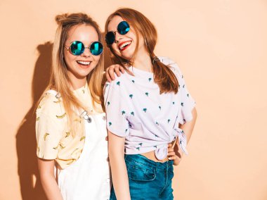 İki genç trendy sarışın hipster kızlarda gülümseyen güzel renkli tişört elbise yaz. Seksi kaygısız kadın yuvarlak güneş gözlüğü bej duvarının yakınında poz. Olumlu modelleri eğleniyor