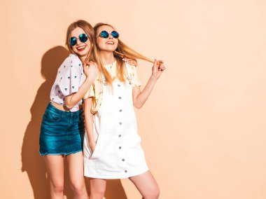 İki genç trendy sarışın hipster kızlarda gülümseyen güzel renkli tişört elbise yaz. Seksi kaygısız kadın yuvarlak güneş gözlüğü bej duvarının yakınında poz. Olumlu modelleri eğleniyor
