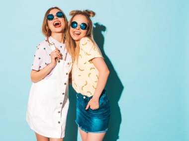 İki genç trendy sarışın hipster kızlarda gülümseyen güzel renkli tişört elbise yaz. Seksi kaygısız kadın yuvarlak güneş gözlüğü mavi duvarının yakınında poz. Olumlu modelleri eğleniyor 