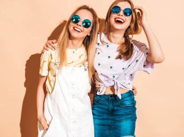 İki genç trendy sarışın hipster kızlarda gülümseyen güzel renkli tişört elbise yaz. Seksi kaygısız kadın yuvarlak güneş gözlüğü bej duvarının yakınında poz. Olumlu modelleri eğleniyor