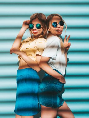 İki genç trendy sarışın hipster kızlarda gülümseyen güzel renkli tişört elbise yaz. Seksi kaygısız kadın yuvarlak güneş gözlüğü mavi duvarının yakınında poz. Eğleniyor ve dilini gösteren pozitif modelleri