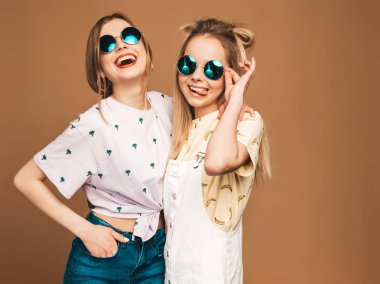 İki genç trendy sarışın hipster kızlarda gülümseyen güzel renkli tişört elbise yaz. Seksi kaygısız kadın yuvarlak güneş gözlüğü bej duvarının yakınında poz. Olumlu modelleri eğleniyor