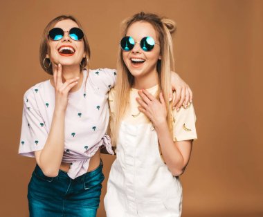 İki genç trendy sarışın hipster kızlarda gülümseyen güzel renkli tişört elbise yaz. Seksi kaygısız kadın yuvarlak güneş gözlüğü bej duvarının yakınında poz. Olumlu modelleri eğleniyor