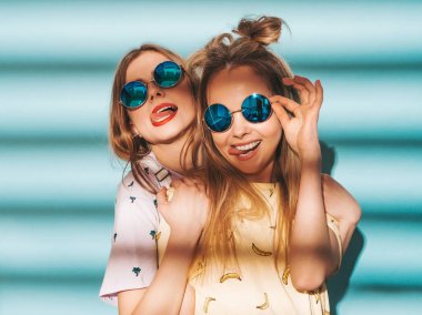 İki genç trendy sarışın hipster kızlarda gülümseyen güzel renkli tişört elbise yaz. Seksi kaygısız kadın yuvarlak güneş gözlüğü mavi duvarının yakınında poz. Eğleniyor ve dilini gösteren pozitif modelleri 
