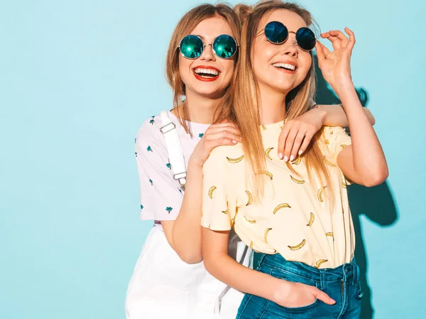 两个年轻美丽的微笑的金发嬉皮士女孩在时尚夏天五颜六色的 T恤衣服 性感无忧无虑的妇女摆出近蓝色墙壁在圆形太阳镜 有乐趣的积极的模型 — 图库照片