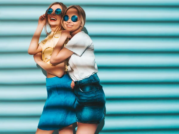 两个年轻美丽的微笑的金发嬉皮士女孩在时尚夏天五颜六色的 T恤衣服 性感无忧无虑的妇女摆出近蓝色墙壁在圆形太阳镜 有乐趣的积极的模型 — 图库照片