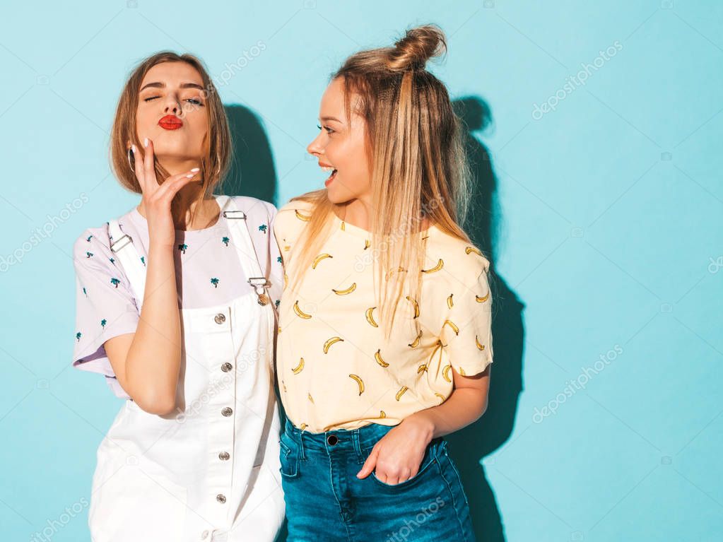 Two young beautiful smiling blond hipster girls in trendy summer colorful T-shirt clothes. Sexy carefree women posing near blue wall. Positive models having fun and giving kiss