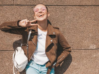 Yaz hipster ceket ve kot elbise giymiş güzel gülümseyen esmer model Portresi. Duvara yakın sokakta poz veren modaya uygun kız. Komik ve olumlu kadın barış işareti gösteren
