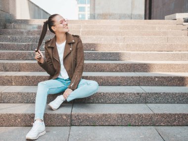 Yaz hipster ceket ve kot elbise giymiş güzel gülümseyen esmer model Portresi. Sokak arka planda merdivenlerde oturan trendy kız. Yuvarlak güneş gözlüğü komik ve pozitif kadın