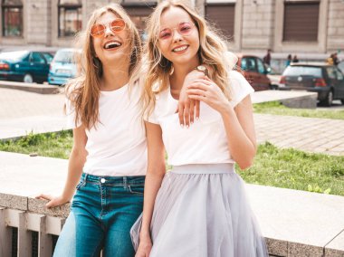 Moda yaz beyaz t-shirt giysiler iki genç güzel sarışın gülümseyen hipster kız Portresi. Seksi kaygısız kadınlar sokak arka planda poz. Güneş gözlüğü ile eğlenen pozitif modeller. Sarılma
