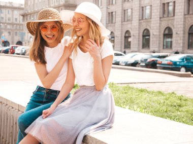 Moda yaz beyaz t-shirt giysiler iki genç güzel sarışın gülümseyen hipster kız Portresi. Seksi kaygısız kadınlar sokak arka planda poz. Güneş gözlüğü ve şapka ile eğlenen pozitif modeller