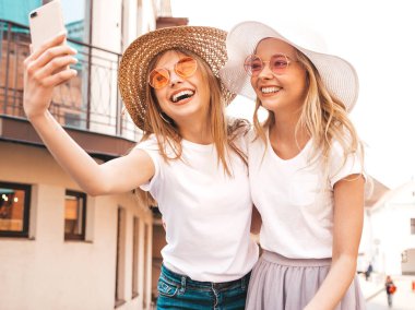 İki genç, gülümseyen, beyaz tişörtlü sarışın kadın. Akıllı telefondan selfie çeken kızlar. Arka planda poz veren mankenler. Olumlu yüz ifadeleri gösteren bir kadın.