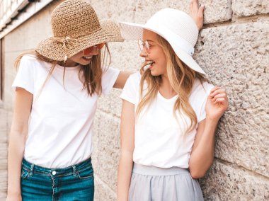 Moda yaz beyaz t-shirt giysiler iki genç güzel sarışın gülümseyen hipster kız Portresi. Seksi kaygısız kadınlar duvara yakın sokakta poz. Güneş gözlüğü ve şapka ile eğlenen pozitif modeller
