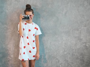 Güzel genç gülümseyen fotoğrafçı kız onu retro kamera kullanarak fotoğraf çekmek. Resim yapan kadın. Model rahat yaz beyaz t-shirt giymiş. Poz Gri duvarın yanındaki stüdyoda