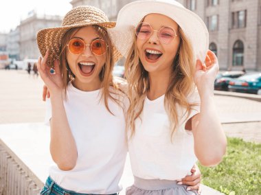 Moda yaz beyaz t-shirt giysiler iki genç güzel sarışın gülümseyen hipster kız Portresi. Seksi kaygısız kadınlar sokak arka planda poz. Güneş gözlüğü ve şapka ile eğlenen pozitif modeller