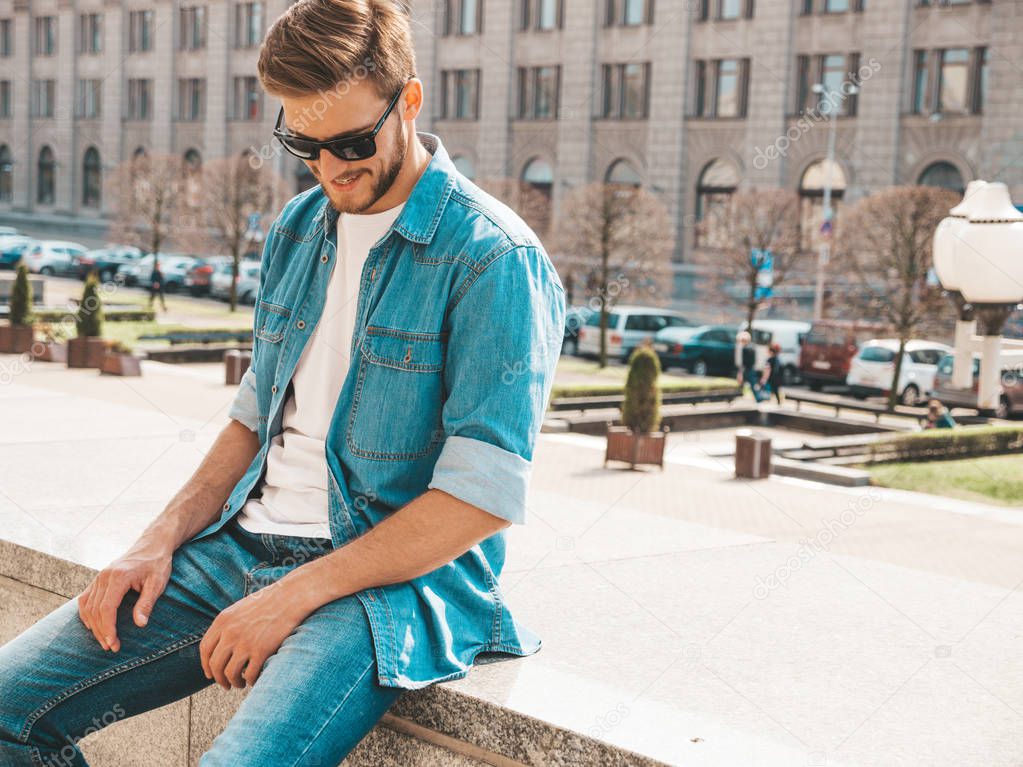 Portrait of handsome smiling stylish hipster lumbersexual businessman model. Man dressed in jeans jacket clothes. Fashion male posing on the street background in sunglasses