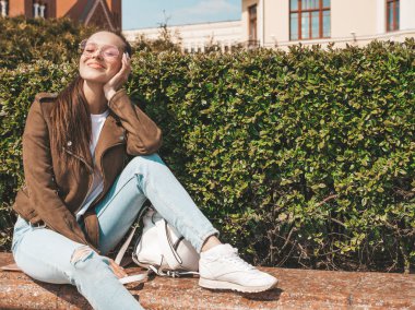 Yaz hipster ceket ve kot elbise giymiş güzel gülümseyen esmer model Portresi. Sokak arka planda bankta oturan trendy kız. Yuvarlak güneş gözlüğü komik ve pozitif kadın