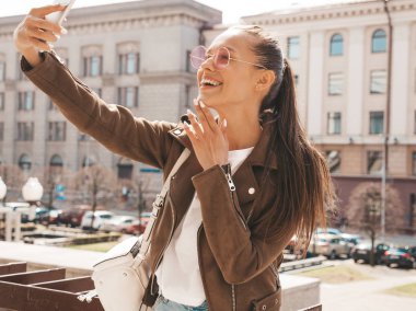 Yaz hipster ceket güzel gülümseyen esmer kız Portresi. Akıllı telefonda selfie çeken model. Güneş gözlüğü ile sokakta sıcak güneşli bir günde fotoğraf yapan kadın