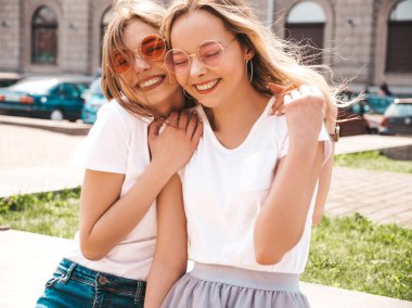 Moda yaz beyaz t-shirt giysiler iki genç güzel sarışın gülümseyen hipster kız Portresi. Seksi kaygısız kadınlar sokak arka planda poz. Güneş gözlüğü ile eğlenen pozitif modeller. Sarılma