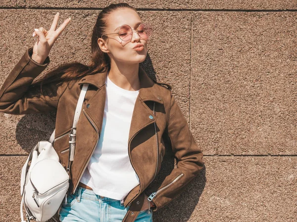 Yaz hipster ceket giysiler giymiş güzel gülümseyen esmer model portresi. Duvara yakın sokakta poz veren modaya uygun kız. Yuvarlak güneş gözlüğü komik ve pozitif kadın. Barış işareti gösterme